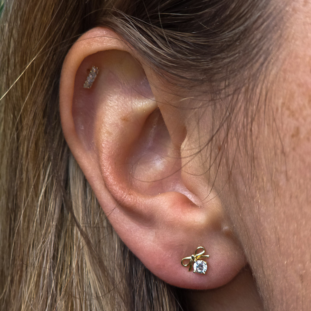 Close-up of an ear wearing a gold bow-shaped earring with a clear gemstone.
