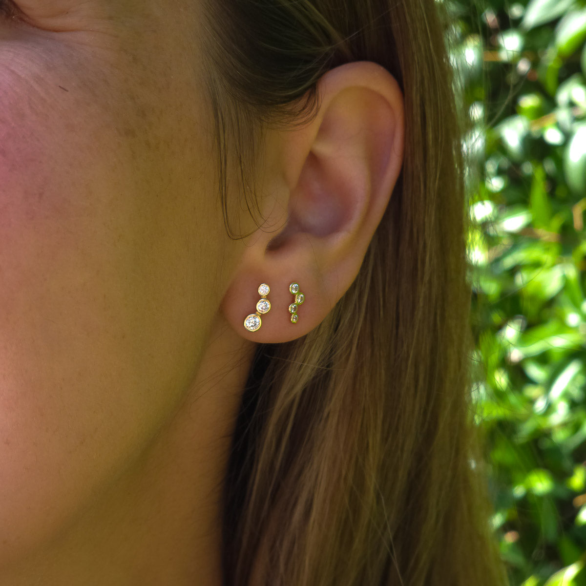 Close-up of an ear wearing gold and diamond stud earrings with a blurred green background