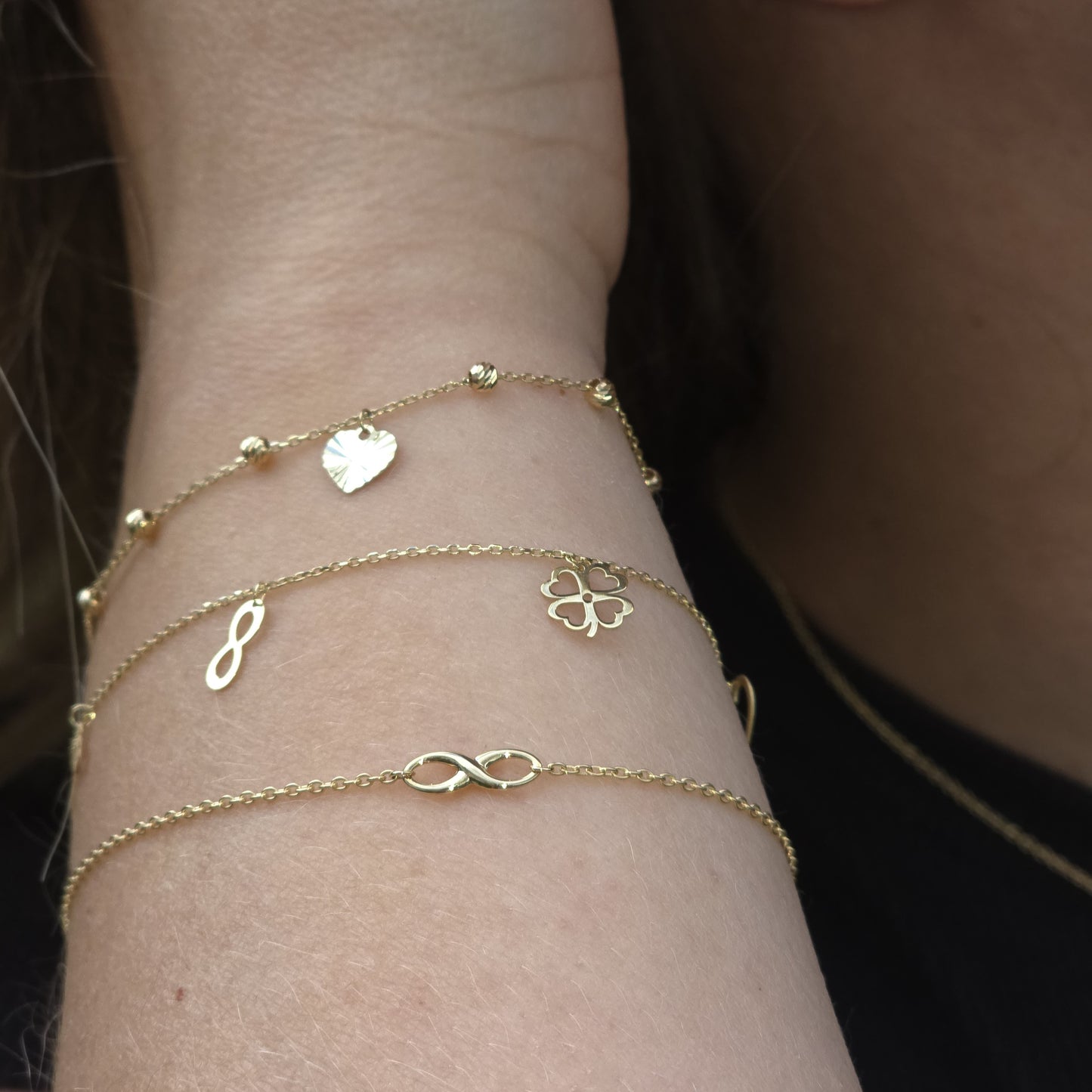 Gold bracelets with heart, infinity, and clover charms on a wrist against a dark background