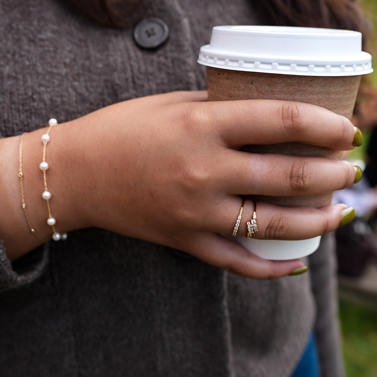 Hand holding a paper cup with a lid, wearing a pearl bracelet and ring, against a blurred background.