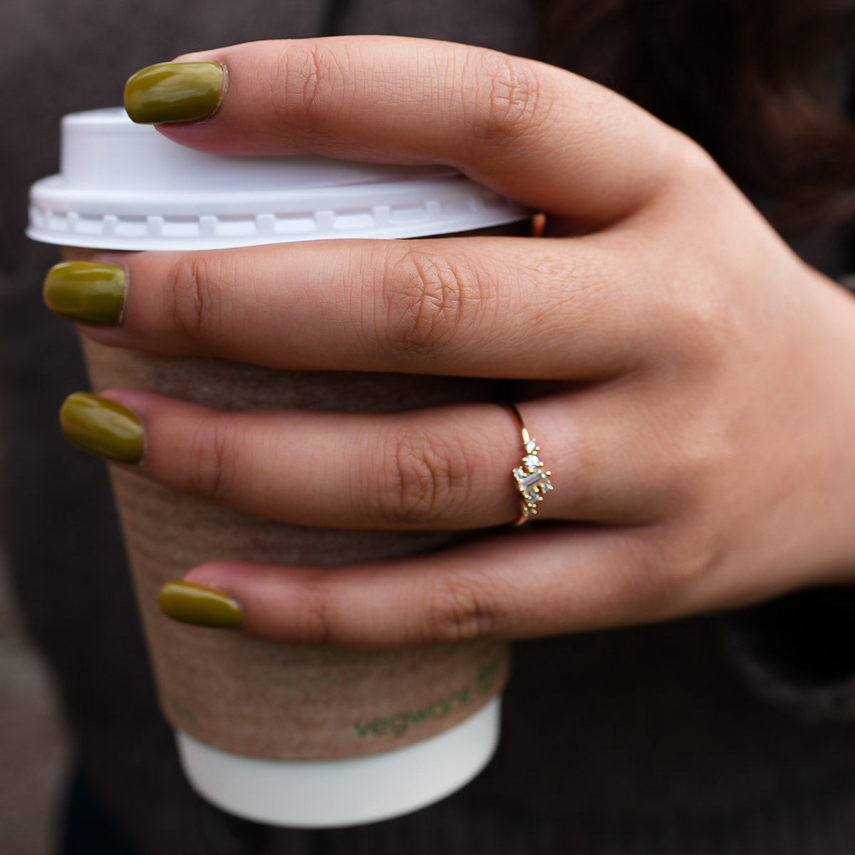 Hand holding a coffee cup with green nail polish and a ring, blurred background