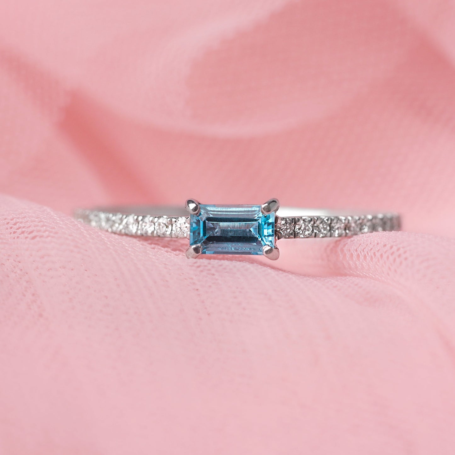Women's solid white gold ring with diamonds adorning the shoulders and a central baguette shape blue topaz gemstone. The ring is displayed on a pale pink tulle background.