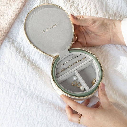 Round jewelry box with 'Stackers' branding, held open to reveal jewelry inside on a white fabric background.
