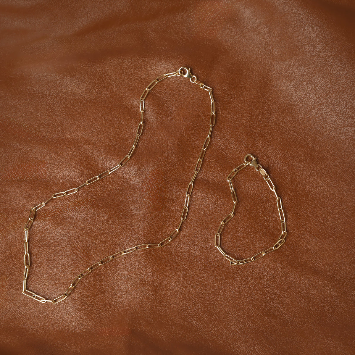 Women's solid yellow gold paperclip chain bracelet and necklace set displayed on a tan brown background.