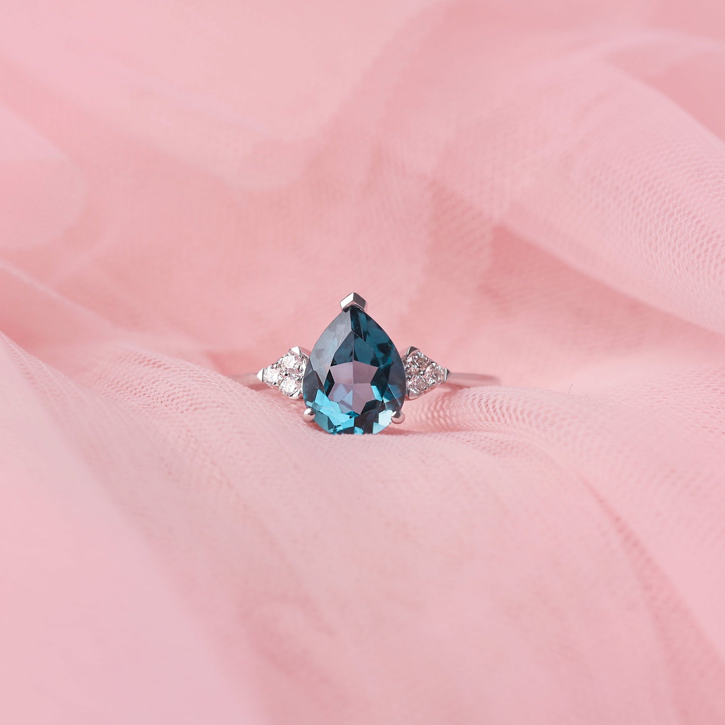 Women's pear shape London blue topaz gemstone claw set on a white gold ring with diamonds framing the centre stone, the ring is displayed on pale pink tulle.