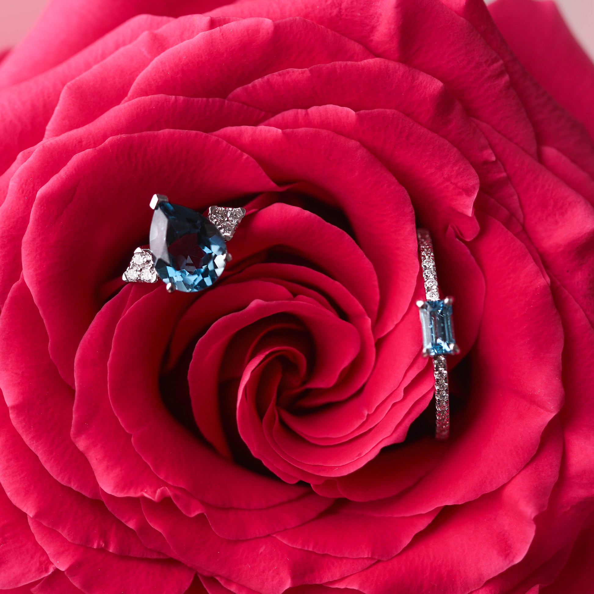 Two women's solid white gold rings set with diamonds on the shoulders, one with a large London blue topaz pear shape gemstone and one with a rectangular blue topaz, all gems are claw set. The rings are displayed on a bright pink rose background.