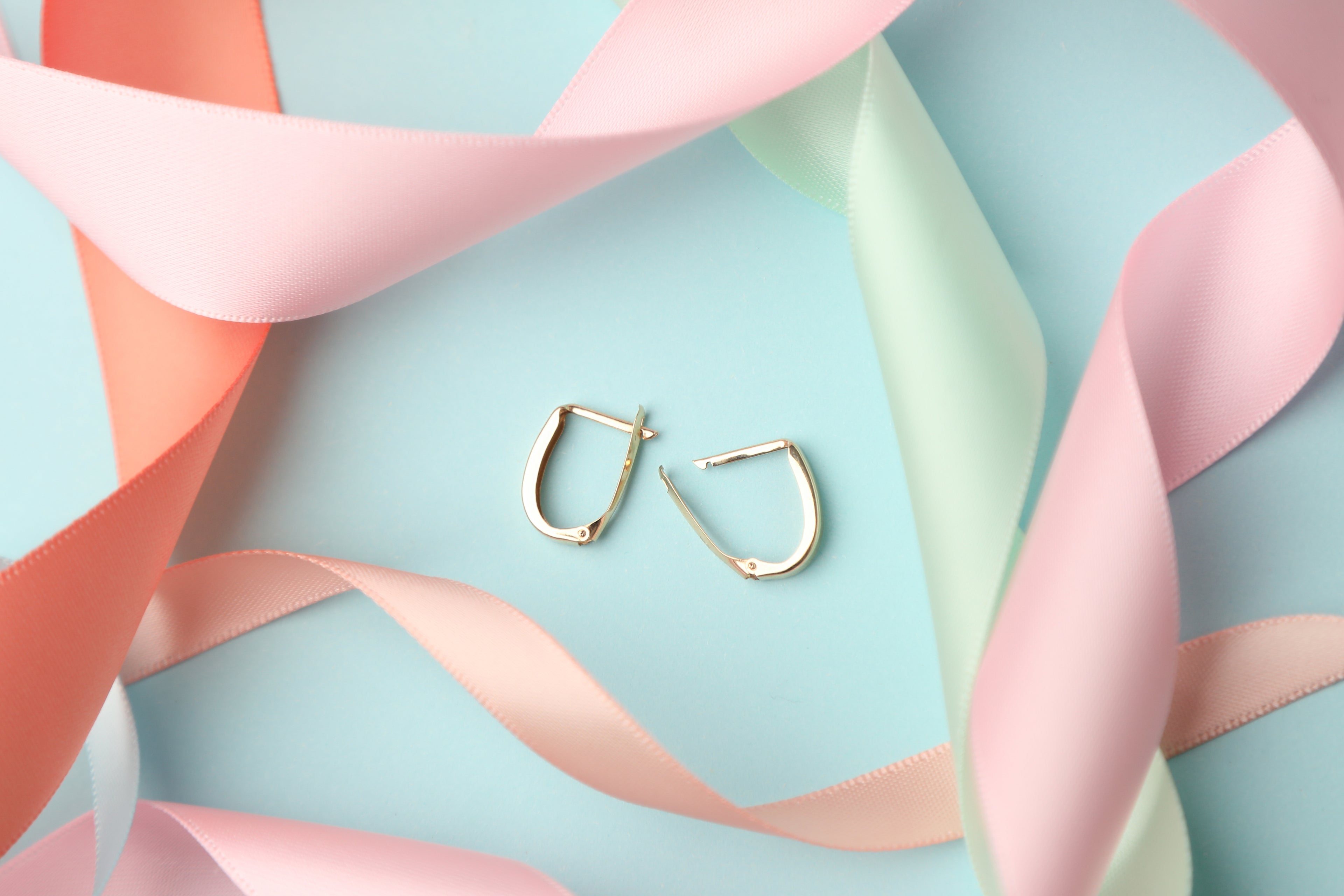 Solid gold paperclip style huggie hoop earrings on a light blue background with pastel ribbons