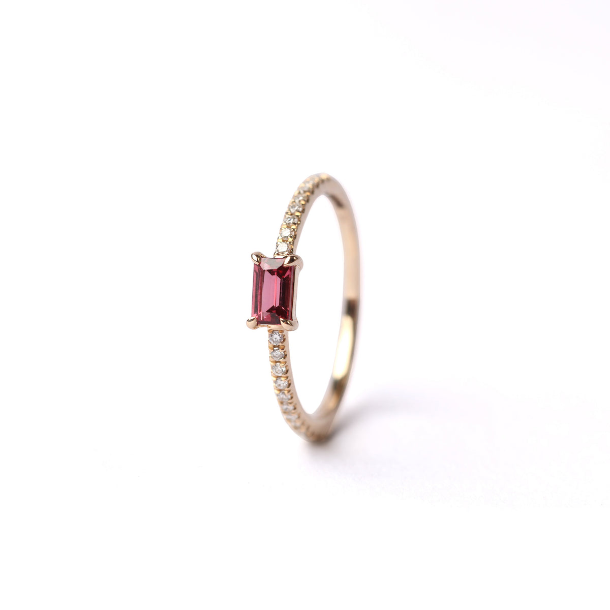 Women's solid yellow gold ring set with a baguette cut pink tourmaline gemstone, with small round diamonds on either side of the shoulders of the ring, displayed standing up, front facing on a white background.