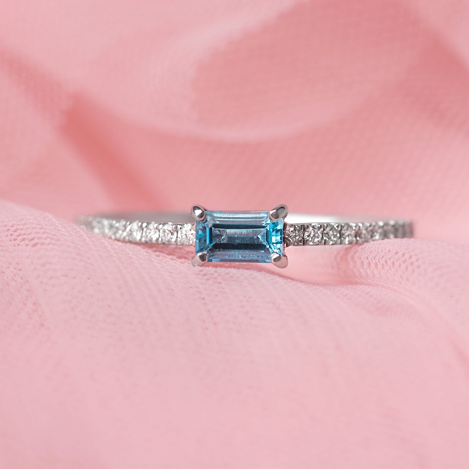 Women's solid white gold ring with diamonds adorning the shoulders and a central baguette shape blue topaz gemstone. The ring is displayed on a pale pink tulle background.