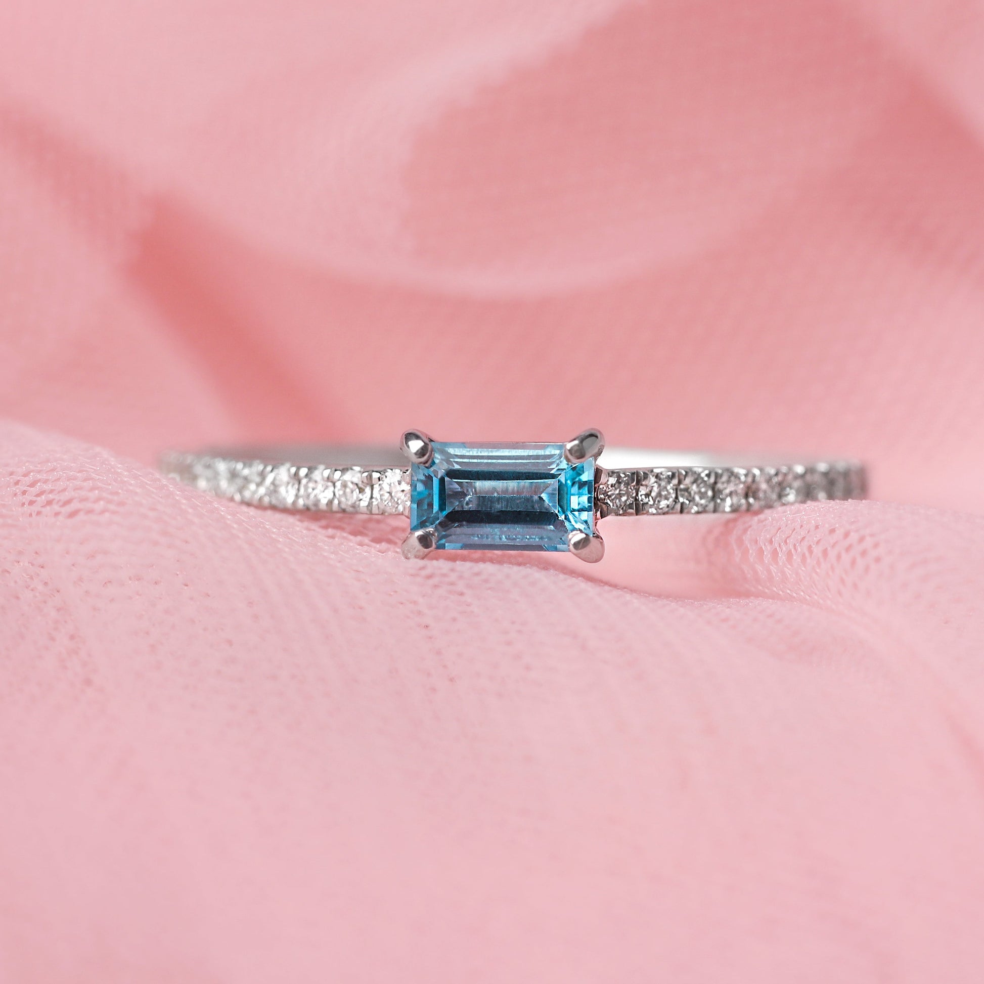Women's solid white gold ring with diamonds adorning the shoulders and a central baguette shape blue topaz gemstone. The ring is displayed on a pale pink tulle background.