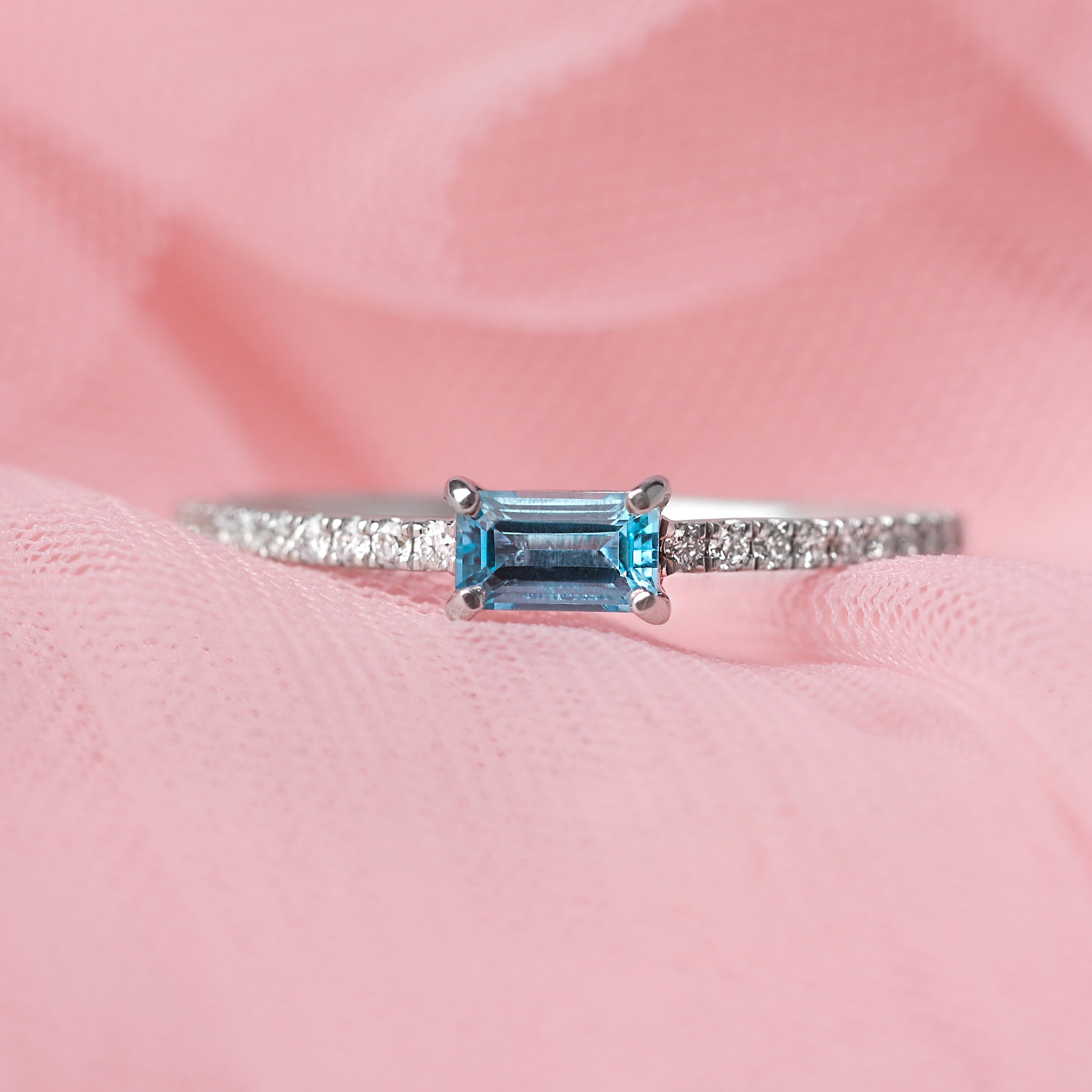 Women's solid white gold ring with diamonds adorning the shoulders and a central baguette shape blue topaz gemstone. The ring is displayed on a pale pink tulle background.