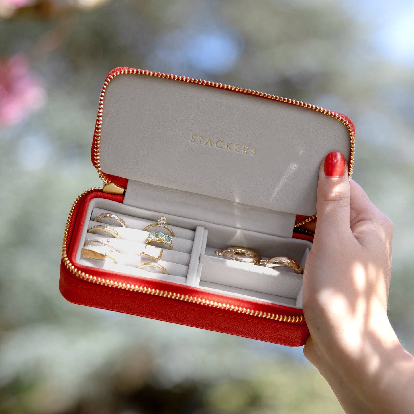 Medium red travel jewellery case with gold rings held by a hand against a blurred natural background