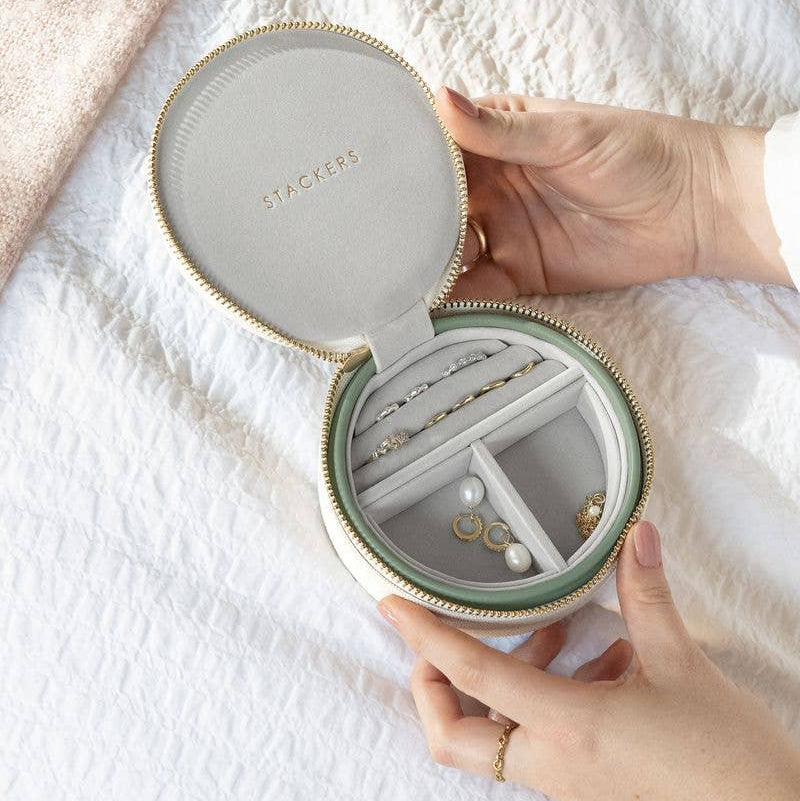Round jewelry box with 'Stackers' branding, held open to reveal jewelry inside on a white fabric background.