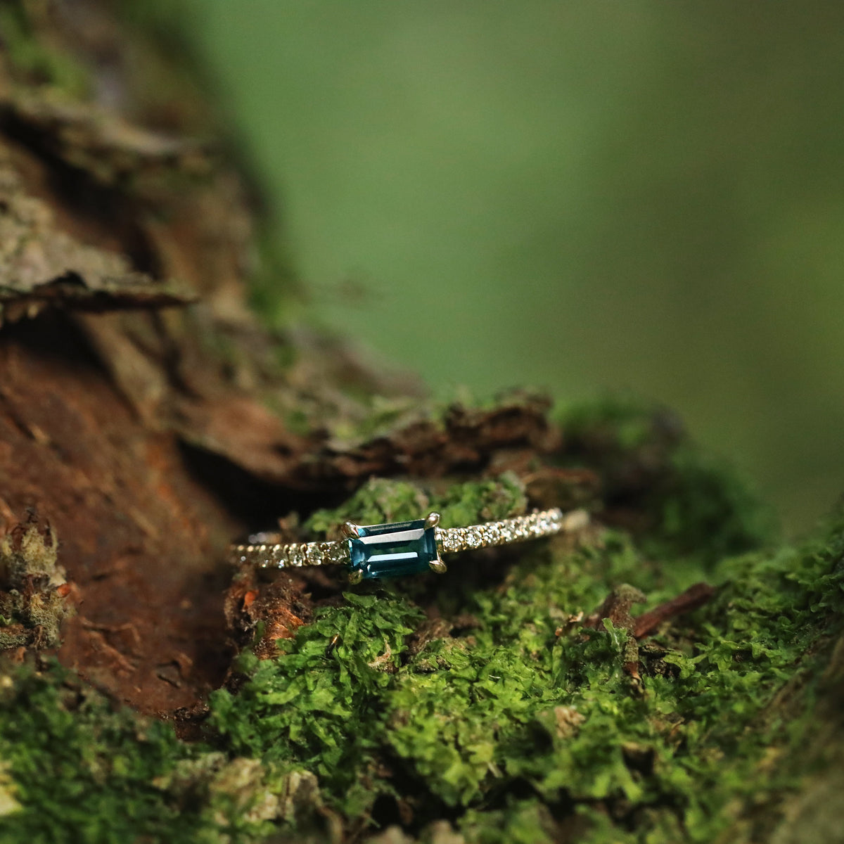A women's solid yellow gold ring set with a baguette cut London blue topaz gemstone placed centrally and small circular diamonds on the shoulders of the ring, displayed on lichen and moss covered tree bark.