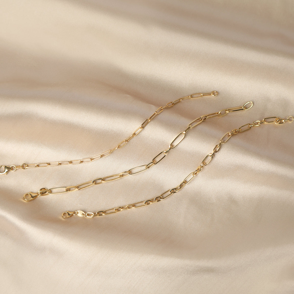 Three yellow gold bracelets made in the paperclip style displayed on a pale champagne gold satin background.
