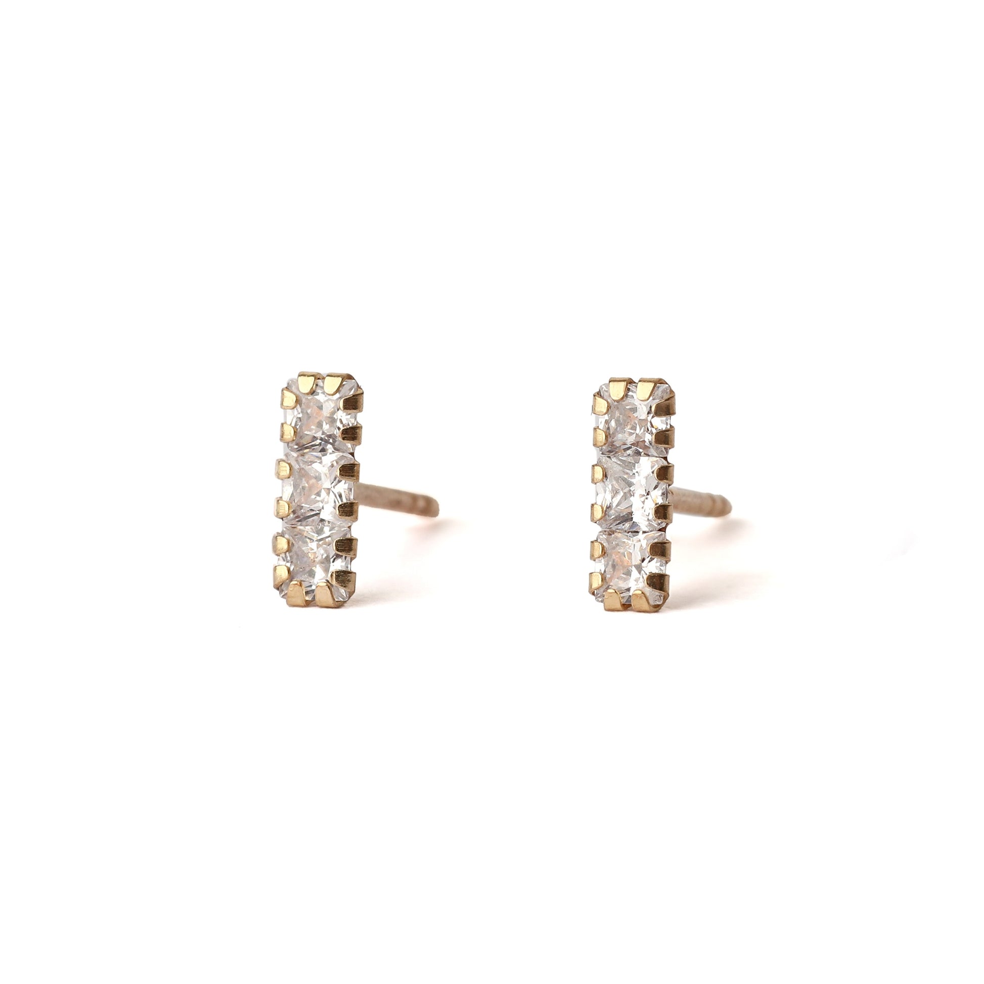 A pair of solid yellow gold studs with three small cubic zirconia gemstones in each, set in a rectangle shape. The earrings dace forward and are displayed for the shopper on a white background.