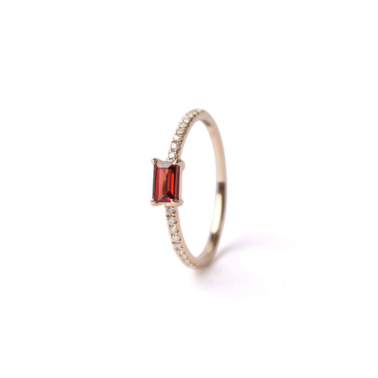 Women's solid yellow gold ring set with diamonds on the shoulders and a central red baguette cut garnet gemstone, displayed on it's side, front facing, on a white background.