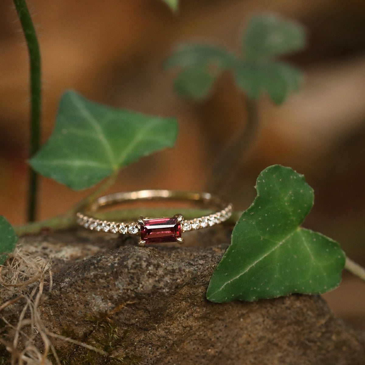 A women's solid yellow gold ring with a rectangular red garnet gemstone and small clear diamond gemstones along the band, resting on a mossy rock surrounded by green ivy leaves in a natural outdoor setting.