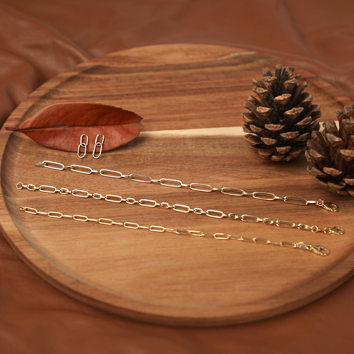 Three yellow gold women's paperclip style chain bracelets and a pair of dangling paperclip style earrings displayed on a wooden board with pine cones and a leaf.