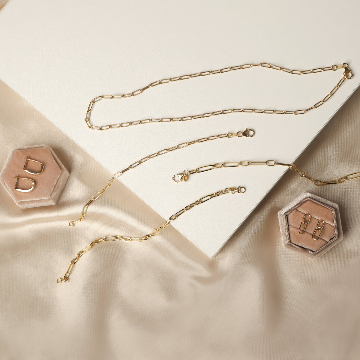 Women's solid yellow gold paperclip chain bracelet and necklace set with two pairs of earrings displayed on a cream beige background.