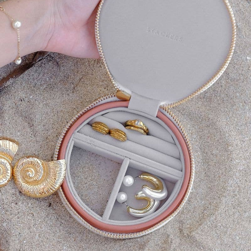 Stackers medium size round travel jewellery box in blush and cream, shown being held by a hand on a sandy surface.