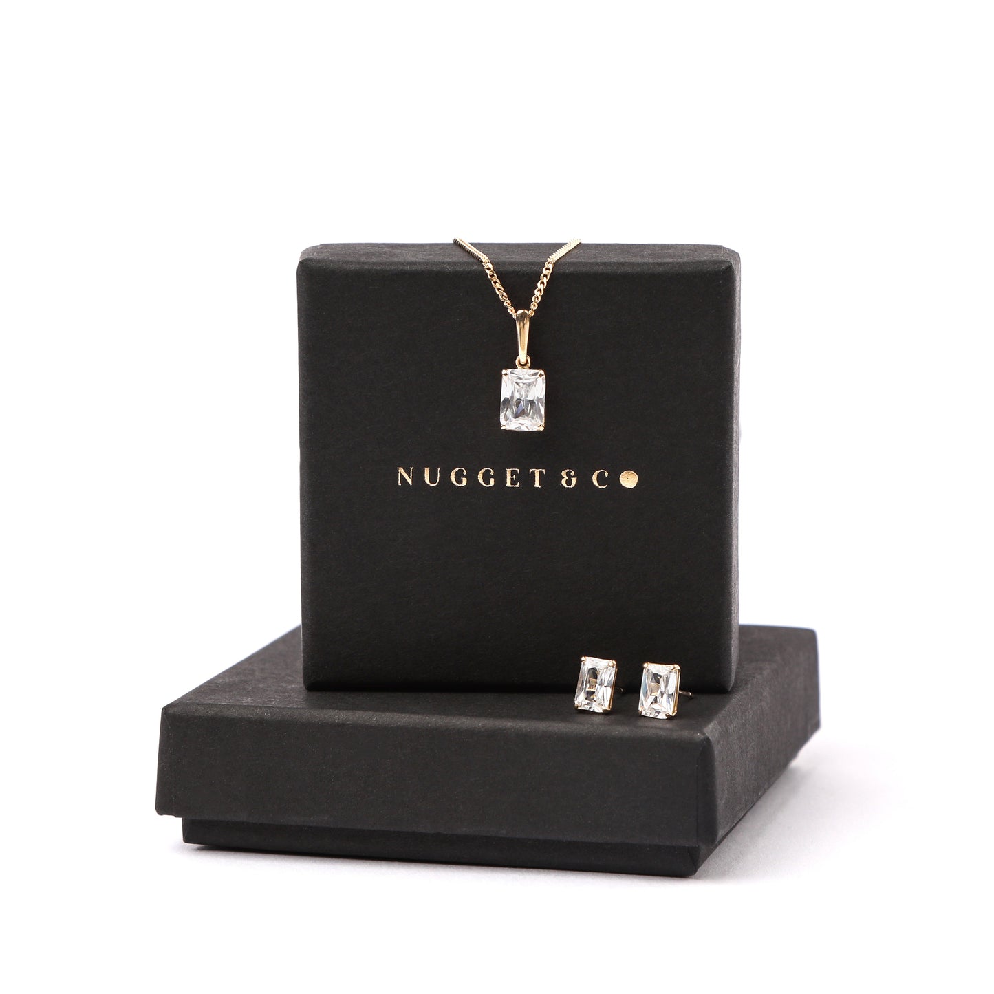 Women's solid gold and emerald-cut cubic zirconia jewellery set with necklace and earrings on a black box labeled 'NUGGET & CO' against a white background.