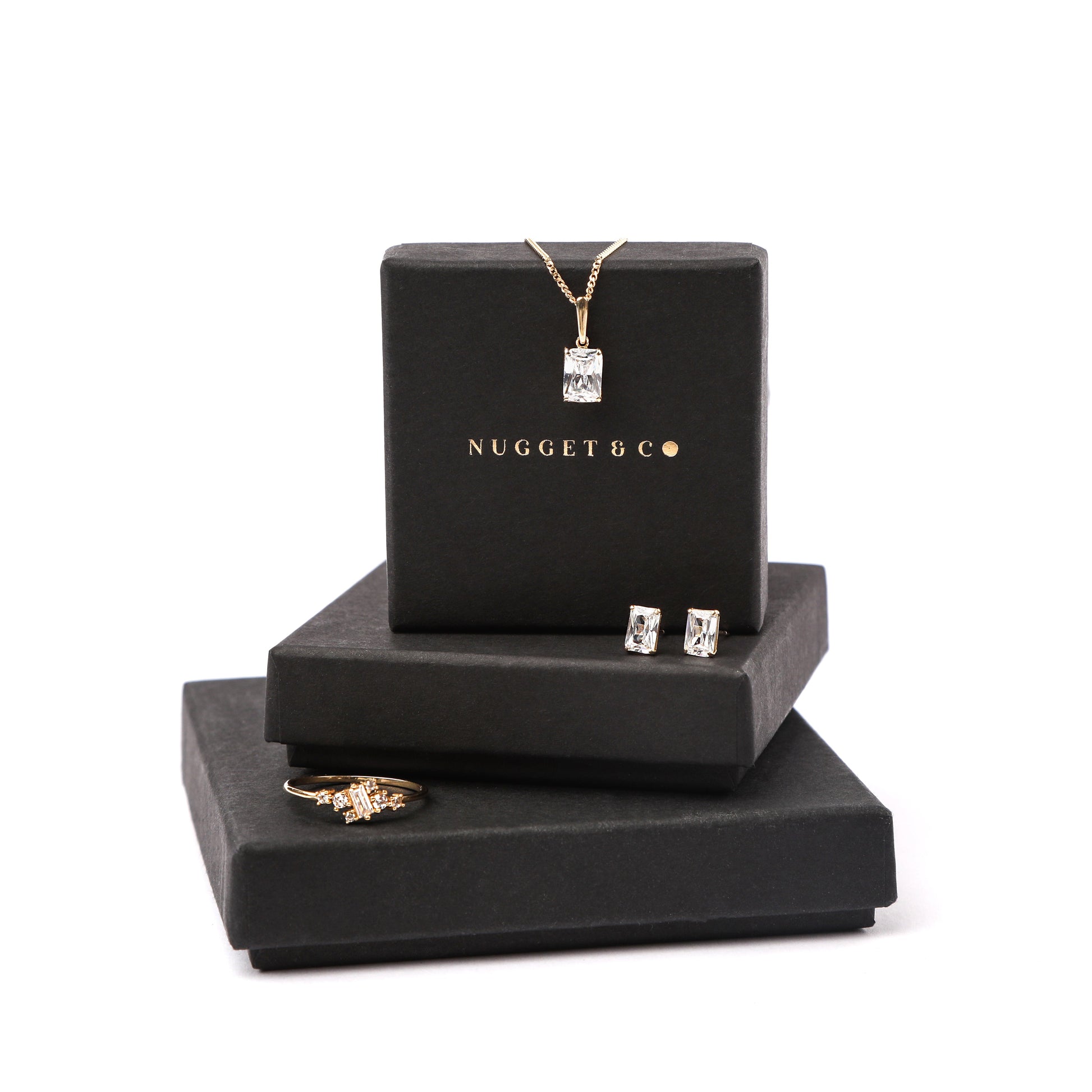 women's solid gold jewellery including emerald-cut cubic zirconia necklace, ring, and earrings on black jewellery boxes with 'Nugget & Co.' branding on a white background.